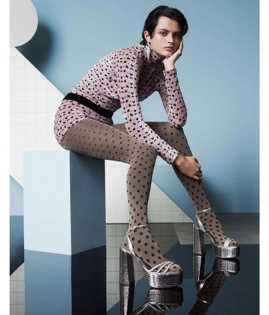 A female model poses indoors wearing a pink patterned long-sleeve shirt and skirt with brown polka-dot tights and chunky silver platform heels.