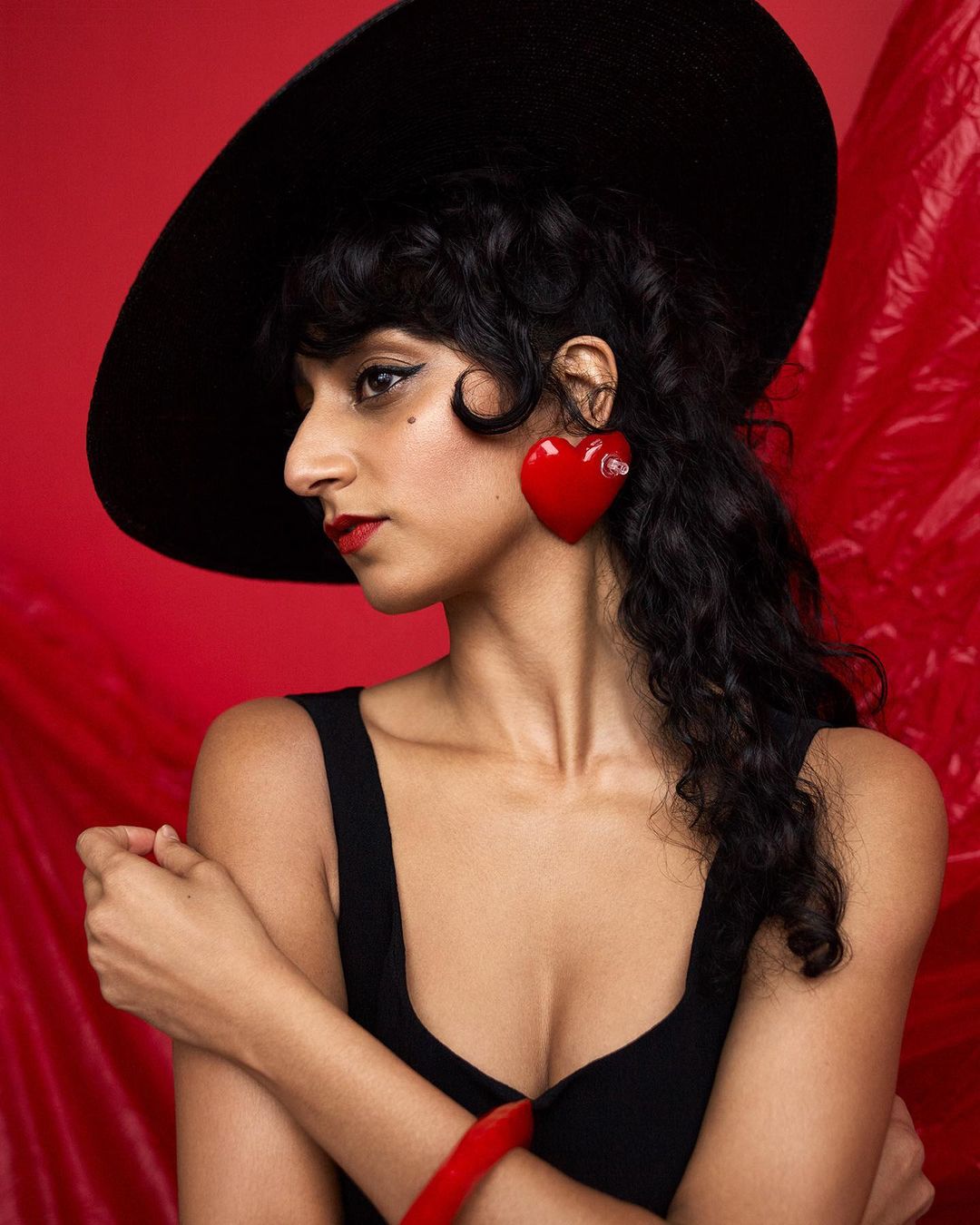 A portrait-style studio shot of a woman with dark curly hair, wearing a black hat and large heart earrings against a red backdrop.