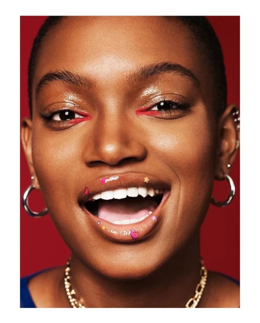 Close-up shot of a smiling adult woman with dark skin, bold red eyeliner, lip art, hoop earrings, and a gold chain against a red backdrop.