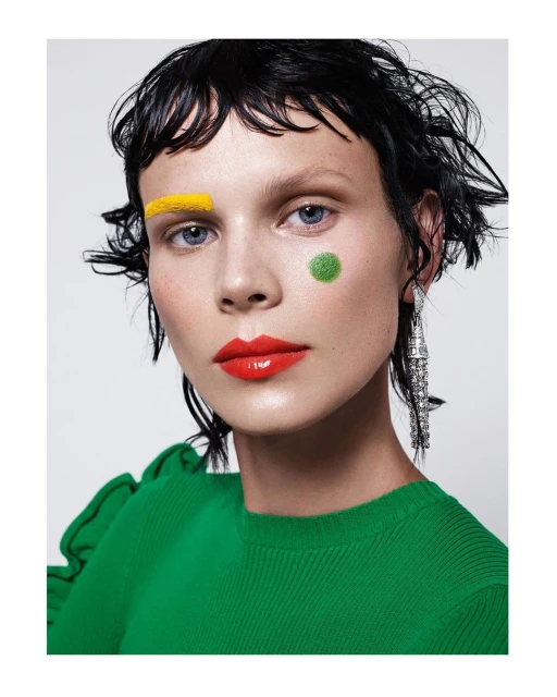 Close-up of a woman with bright yellow, green, and red makeup and glossy red lipstick, accessorized with chandelier earrings against a white backdrop.
