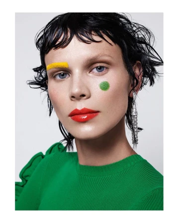 Close-up of a woman with bright yellow, green, and red makeup and glossy red lipstick, accessorized with chandelier earrings against a white backdrop.