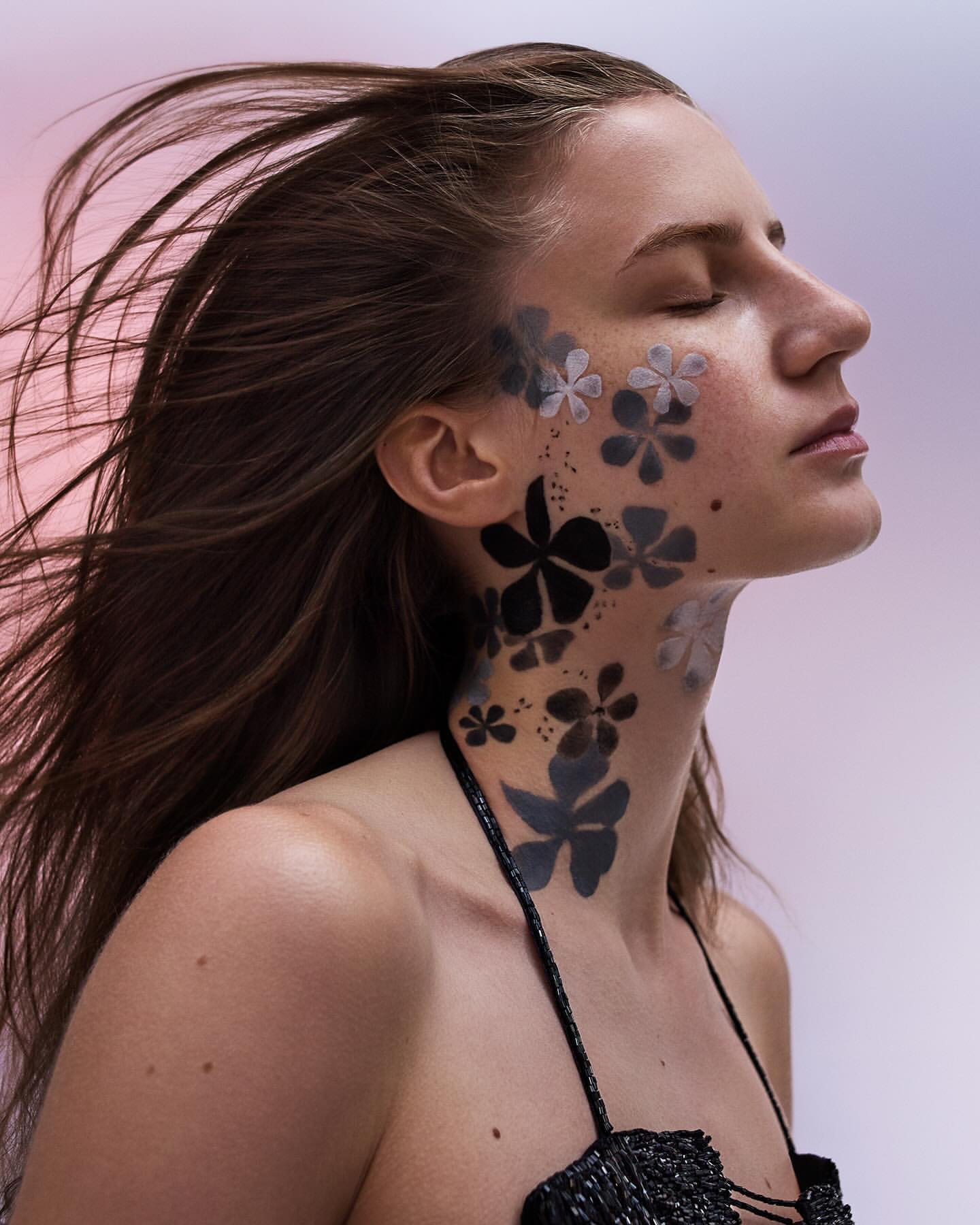Close-up of a woman with wind-blown hair and artistic floral face paint in black and grey, set against a soft pink gradient.