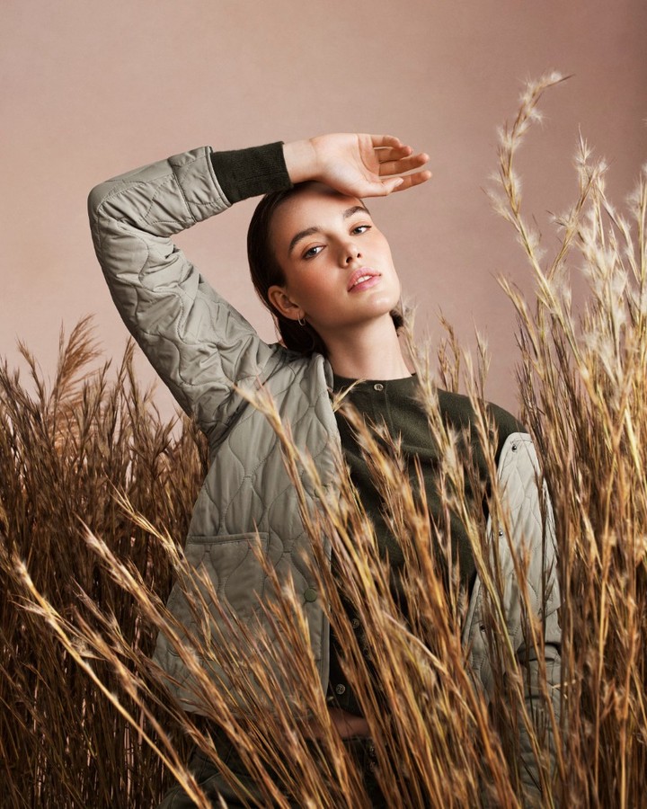 Stylish adult woman poses with hand above brow, wearing a quilted sage jacket in front of light tan background and pampas grass.