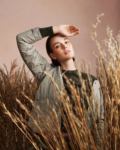 Stylish adult woman poses with hand above brow, wearing a quilted sage jacket in front of light tan background and pampas grass.