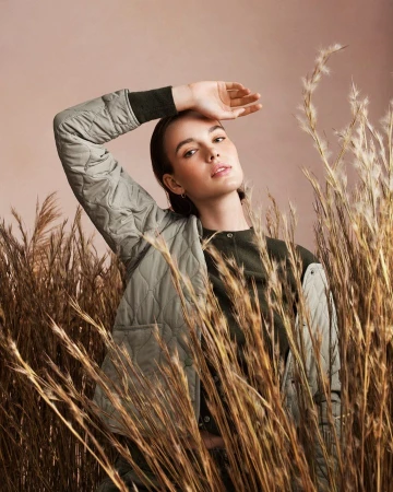 Stylish adult woman poses with hand above brow, wearing a quilted sage jacket in front of light tan background and pampas grass.