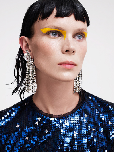 Close up portrait of a woman with vibrant yellow eye makeup, crystal statement earrings, and a sequined blue top.