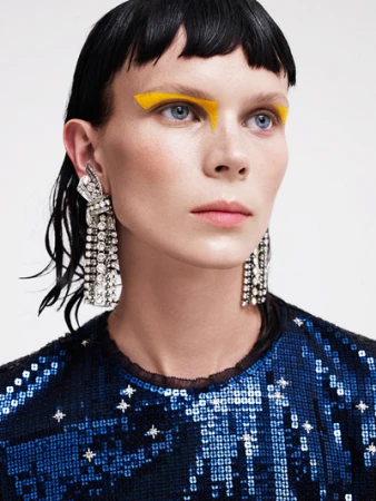 Close up portrait of a woman with vibrant yellow eye makeup, crystal statement earrings, and a sequined blue top.