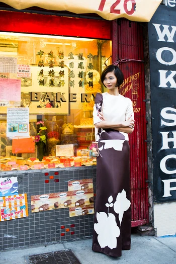 Asian woman in floral dress stands in front of bakery window with desserts, evoking a vintage fashion editorial and urban street style.