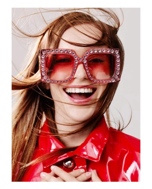 Close-up of a smiling, light-skinned woman wearing large, pink, jeweled sunglasses and a shiny red jacket against a plain background.