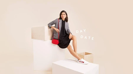 An adult woman poses in a black dress, grey blazer, and white sneakers, sitting on white blocks in a studio with a beige backdrop.