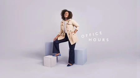 A woman in a trench coat and jeans poses on blocks in a studio with a slight smile, dressed in an outfit for work or a casual outing.
