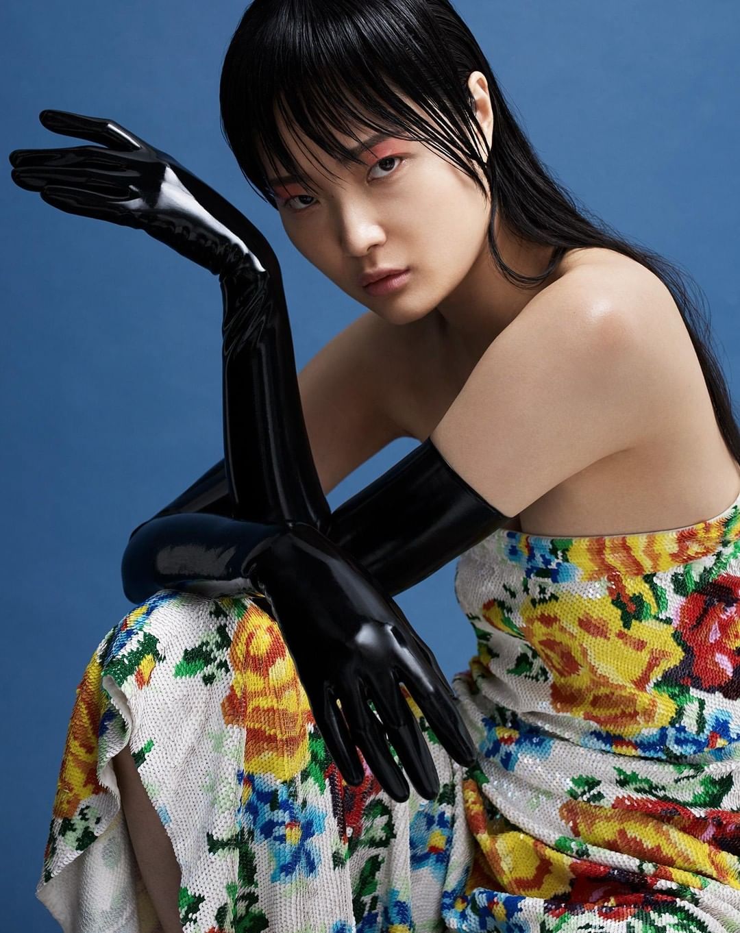 Fashion portrait of a young Asian woman in floral dress and black latex gloves against a blue backdrop, posing with arms raised.