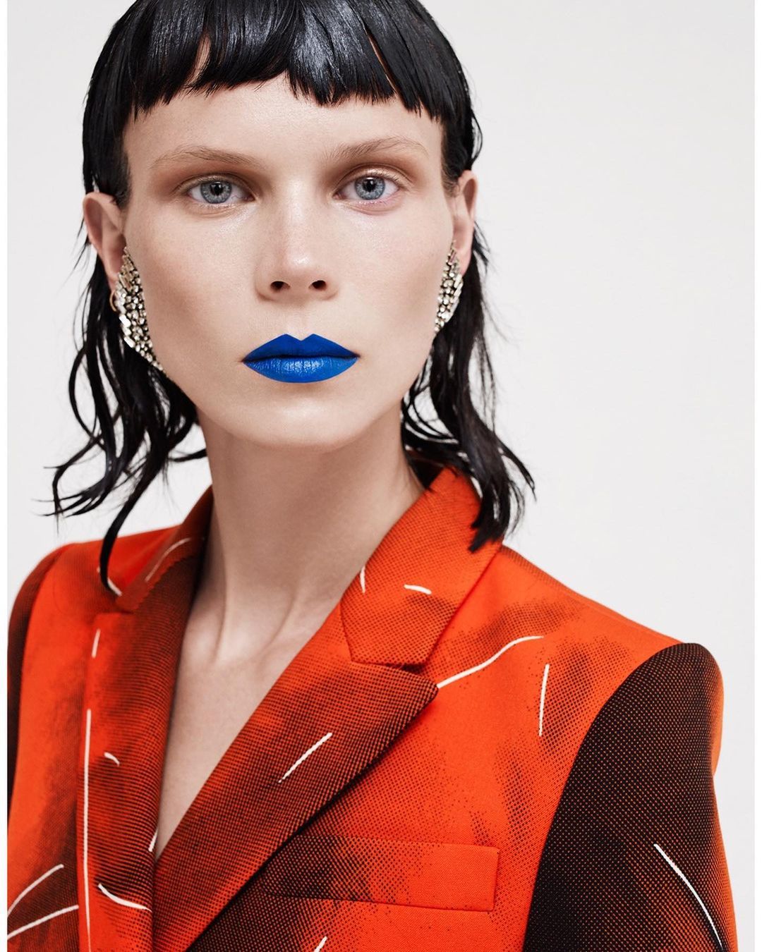 Fashion portrait of a woman with blue lipstick wearing a printed blazer, silver earrings, and a glossy dark fringe against a white backdrop.