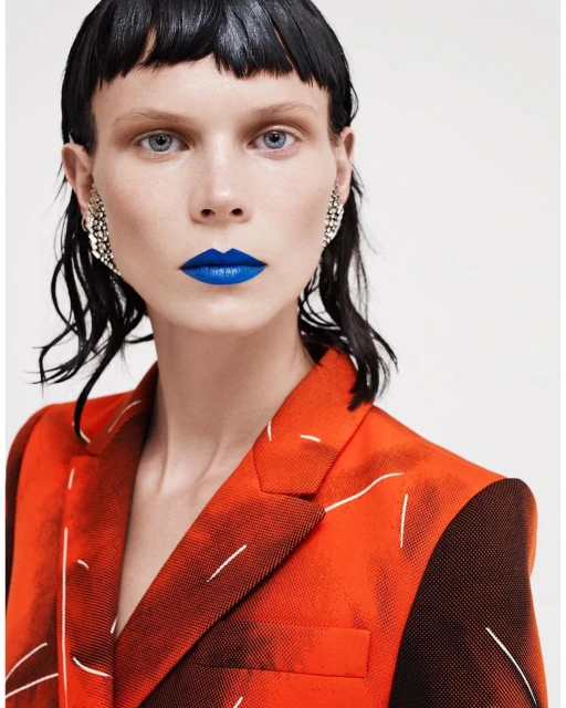 Fashion portrait of a woman with blue lipstick wearing a printed blazer, silver earrings, and a glossy dark fringe against a white backdrop.