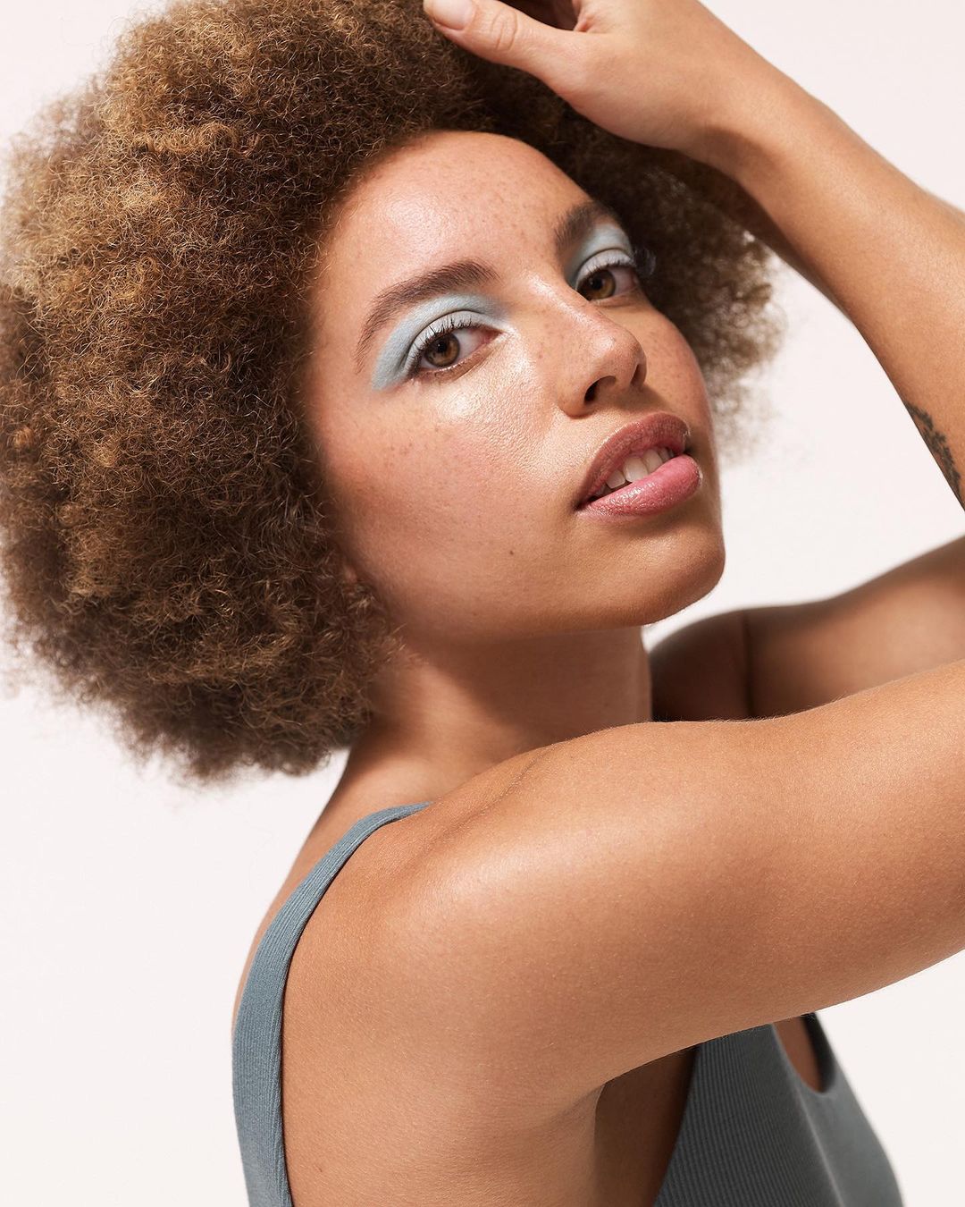 Close-up of an adult female with tan skin and curly brown hair wearing light blue eyeshadow, posing against a light neutral background.