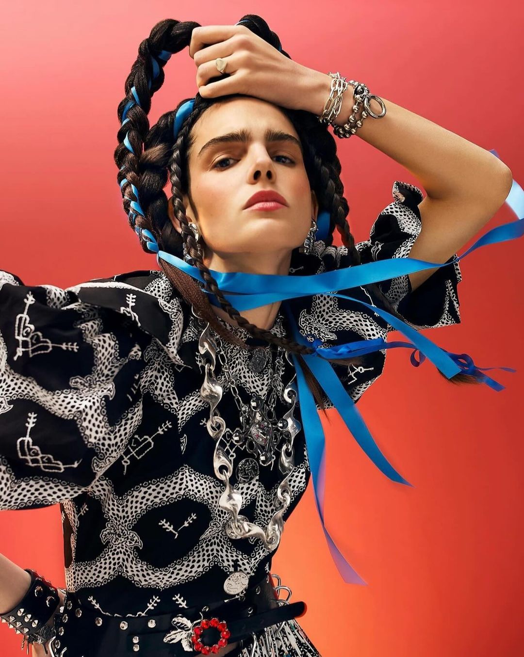 Adult female posing in black patterned shirt, accessorized with silver necklace, bracelets, rings, and braided hair decorated with blue ribbon, in studio.