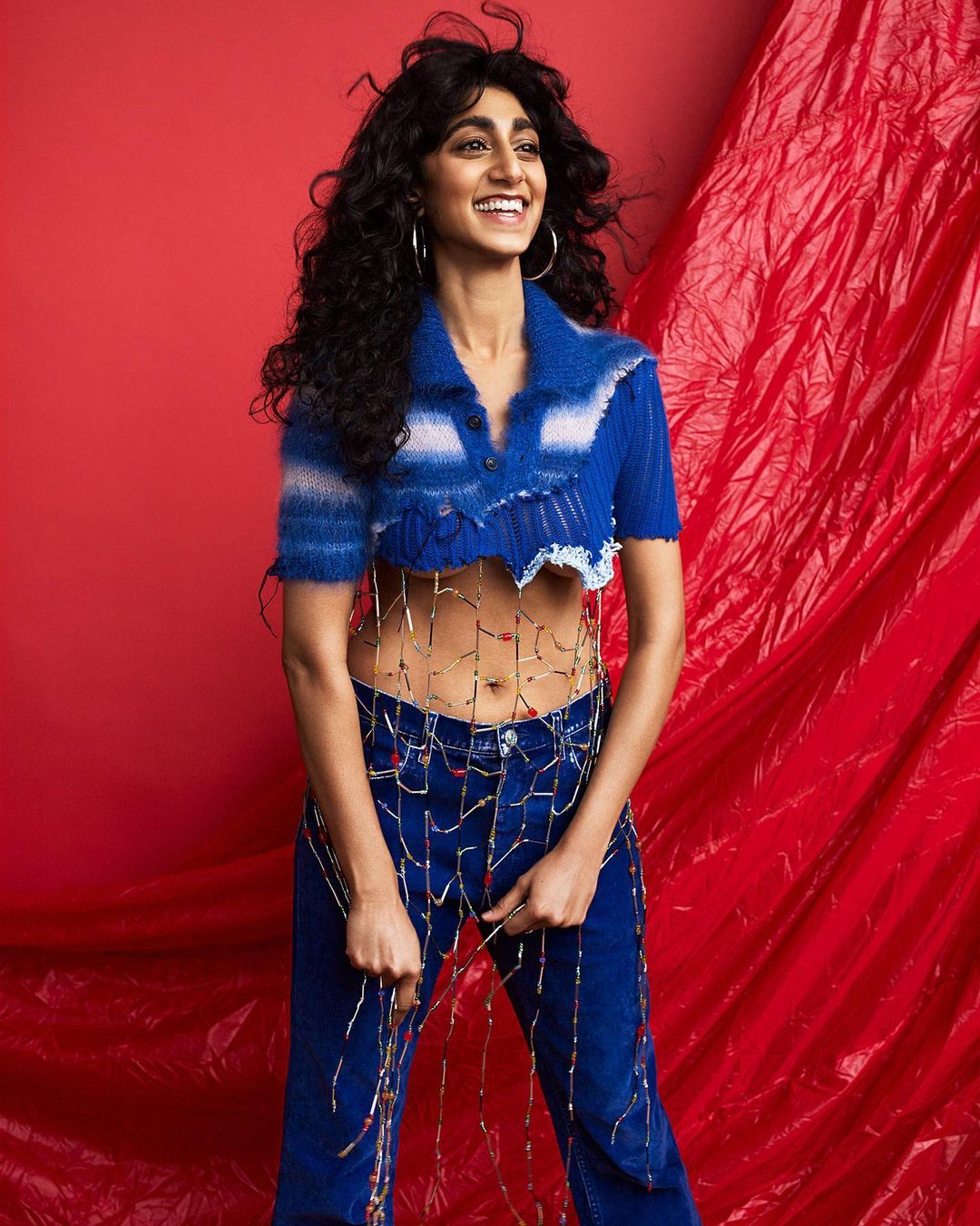 Smiling woman in a blue crop top and jeans posing against a red backdrop with artistic lighting and hair, fashion-forward and stylish.