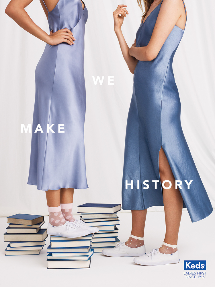 Two women in satin dresses stand on stacks of books, showcasing footwear. The backdrop is a simple white, emphasizing the fashion.