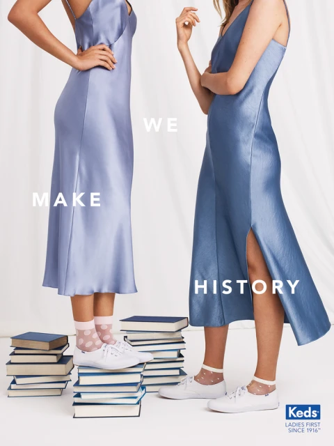 Two women in satin dresses stand on stacks of books, showcasing footwear. The backdrop is a simple white, emphasizing the fashion.