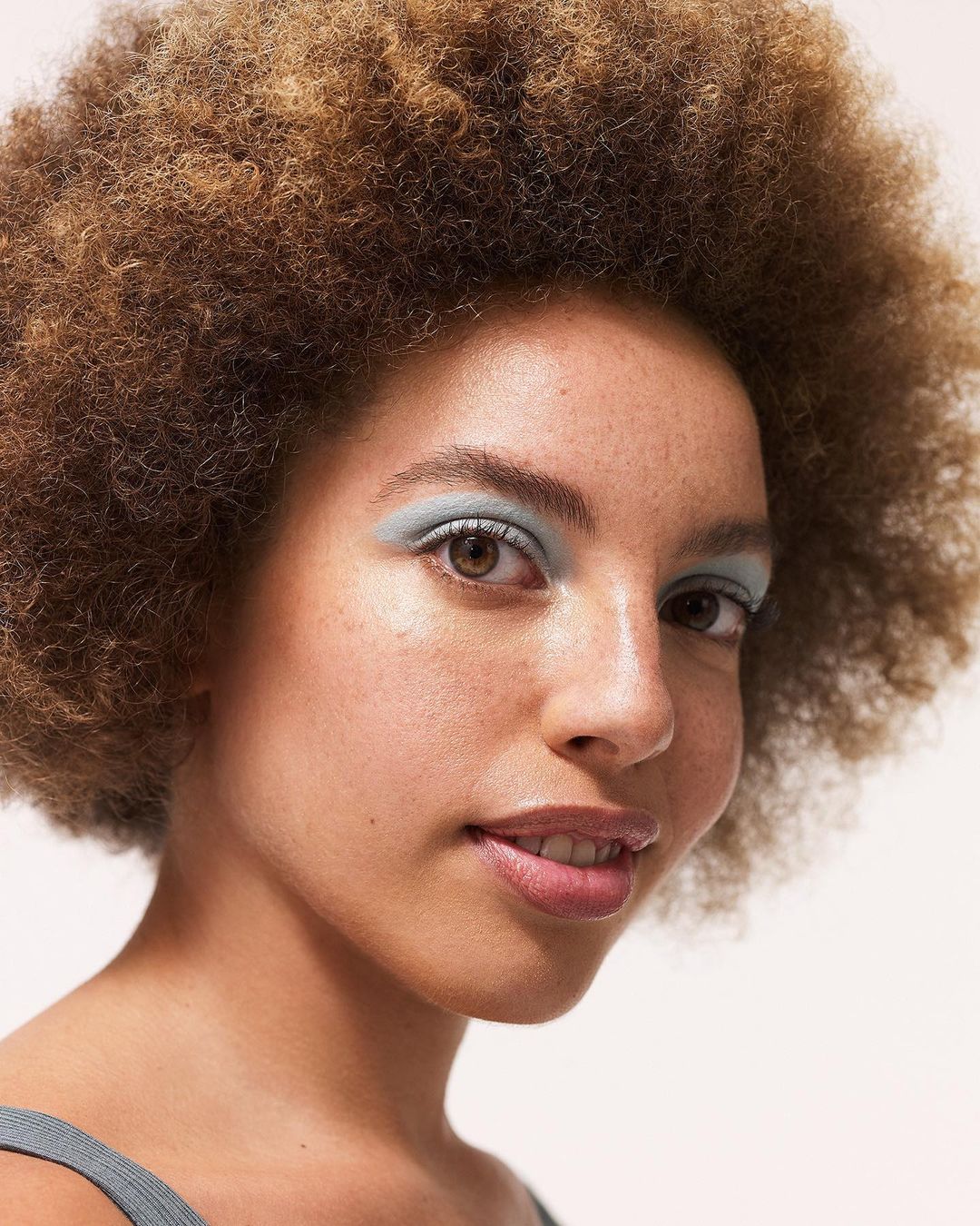 Close-up portrait of a beautiful young woman with an afro, wearing blue eyeshadow, and smiling in a well-lit, neutral indoor setting.
