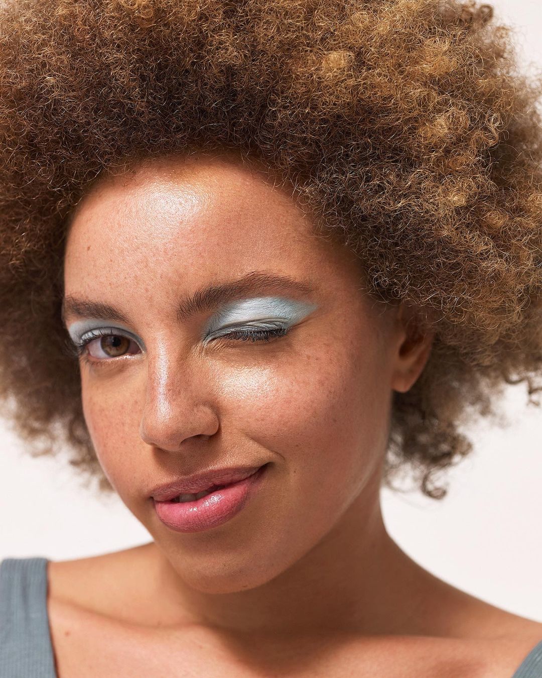 A young adult female with curly brown hair winks at the camera while wearing light blue eyeshadow, in a close-up studio portrait.