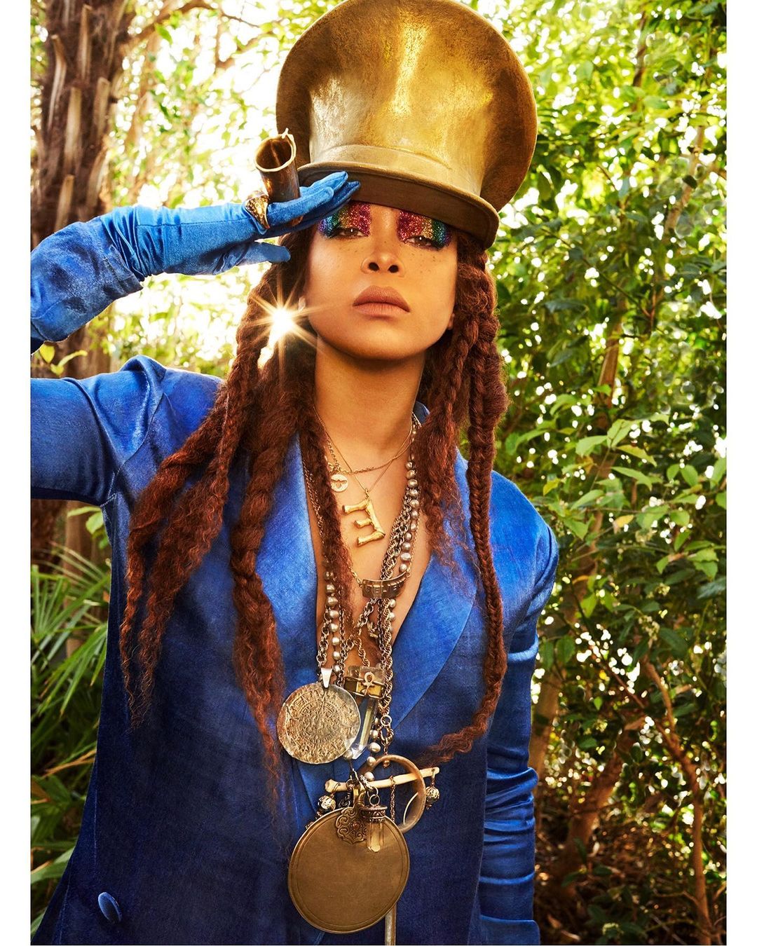 Woman with elaborate eye makeup and large hat poses in a blue suit outdoors surrounded by greenery, looking confident and stylish.