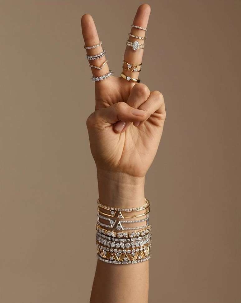 Hand making a peace sign gesture, adorned with multiple rings, with a variety of gold and diamond bracelets visible on the wrist against a neutral background.