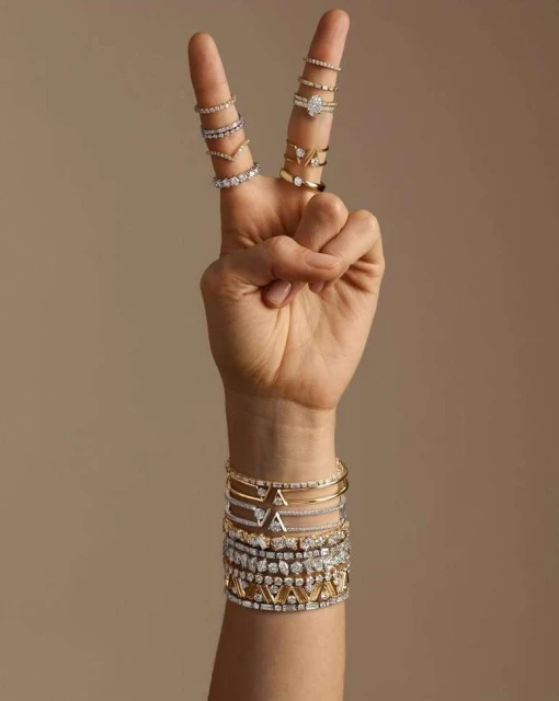 Hand making a peace sign gesture, adorned with multiple rings, with a variety of gold and diamond bracelets visible on the wrist against a neutral background.