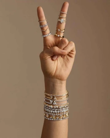 Hand making a peace sign gesture, adorned with multiple rings, with a variety of gold and diamond bracelets visible on the wrist against a neutral background.