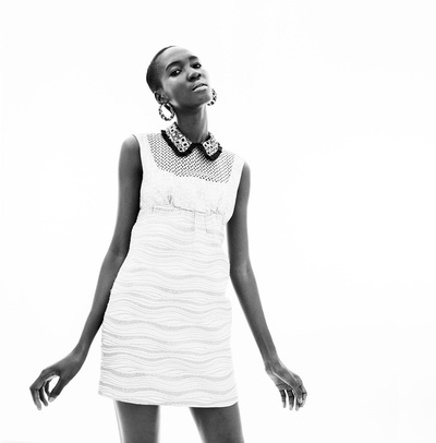 Black and white studio portrait of a young woman in a white dress with a detailed collar and hoop earrings against a white background.