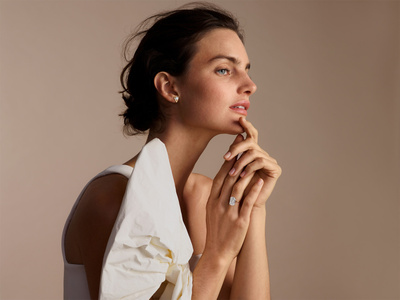 Elegant brunette woman poses in soft light with a diamond ring and off-the-shoulder white top in a beauty and jewelry advertisement.
