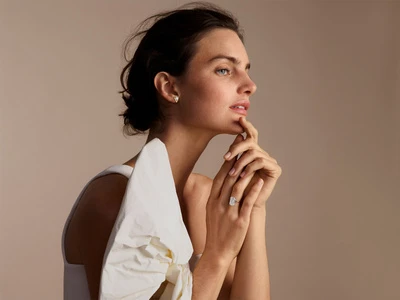 Elegant brunette woman poses in soft light with a diamond ring and off-the-shoulder white top in a beauty and jewelry advertisement.