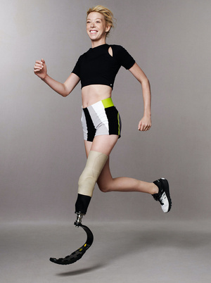 Athletic woman with prosthetic leg smiles while running and jumping mid-air, wearing athletic shorts and a black top against grey backdrop.