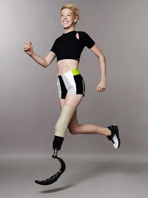Athletic woman with prosthetic leg smiles while running and jumping mid-air, wearing athletic shorts and a black top against grey backdrop.