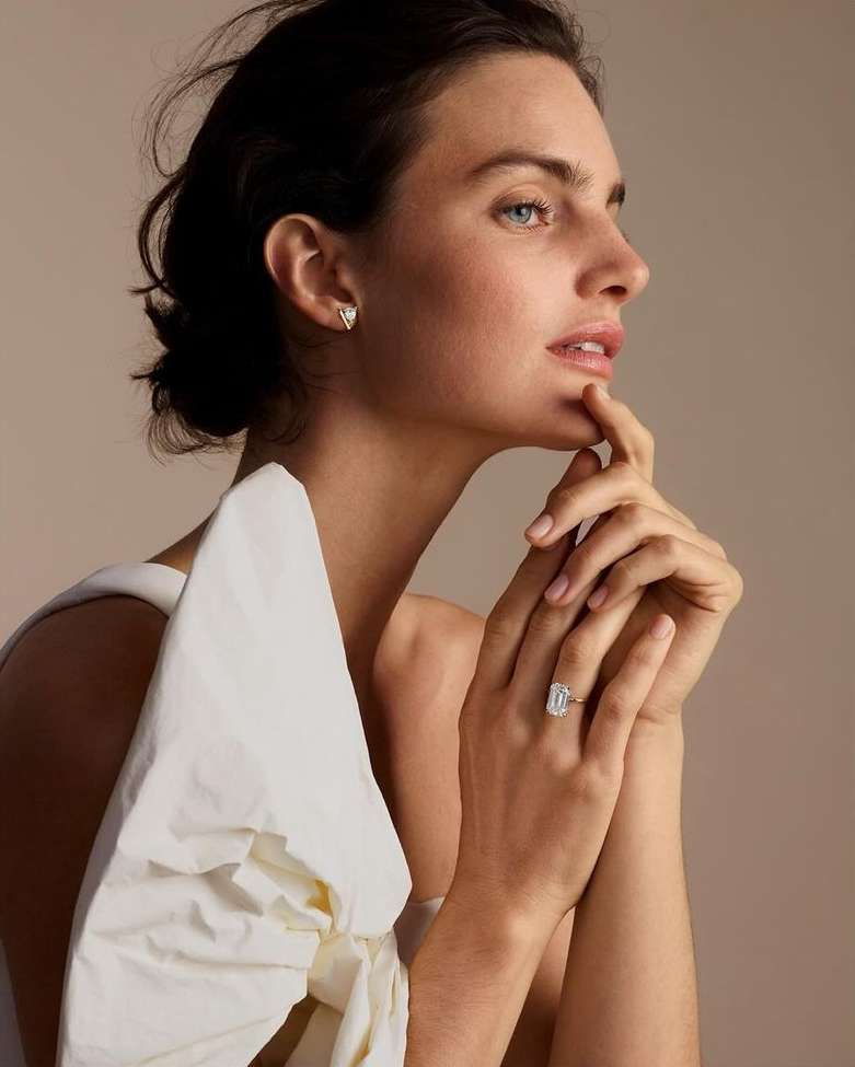 Close-up profile portrait of a woman wearing a large diamond ring and triangular stud earrings against a warm neutral backdrop.