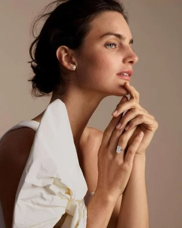 Close-up profile portrait of a woman wearing a large diamond ring and triangular stud earrings against a warm neutral backdrop.