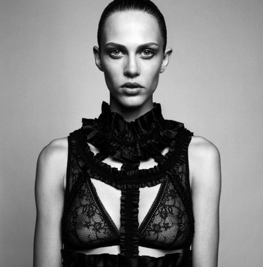 Monochrome portrait of a woman in a lace bra and ruffled high collar, set against a neutral backdrop in a studio environment.