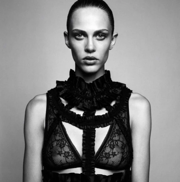 Monochrome portrait of a woman in a lace bra and ruffled high collar, set against a neutral backdrop in a studio environment.