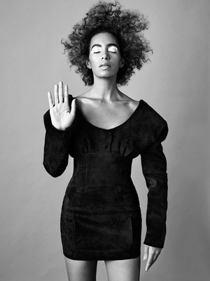 Black and white portrait of an adult woman with curly hair, wearing a black dress with a raised palm, against a neutral background.