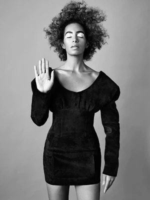 Black and white portrait of an adult woman with curly hair, wearing a black dress with a raised palm, against a neutral background.