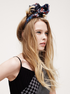 Blonde woman in a black dress and floral headband poses in a studio with soft lighting, creating a fashion portrait.