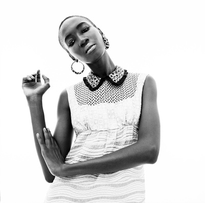 Black and white shot of a Black woman in a sleeveless dress with hoop earrings posing in front of a white backdrop.