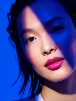 Close-up portrait of an Asian woman with bright pink lips, illuminated by a bar of vivid blue lighting across her face.