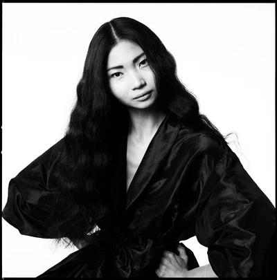 Black and white portrait of an Asian woman with long wavy hair wearing a silk robe against a bright white backdrop in a photo studio.