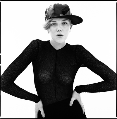 Monochrome portrait of a woman wearing a black long-sleeve mesh top and a dark baseball cap against a white background.