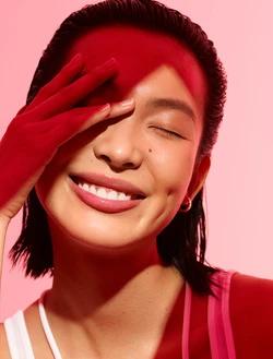 Close-up portrait of a smiling woman with wet-look dark hair holding a red-gloved hand over her forehead against a soft pink background.