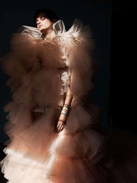 An adult female model poses indoors wearing a layered, ruffled, light peach colored gown with arm jewelry in a fashion photography setting.