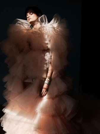 An adult female model poses indoors wearing a layered, ruffled, light peach colored gown with arm jewelry in a fashion photography setting.