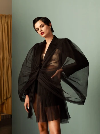 A model poses indoors wearing a sheer black blazer-dress against a muted teal background with gold curtain accent, capturing fashion elegance.