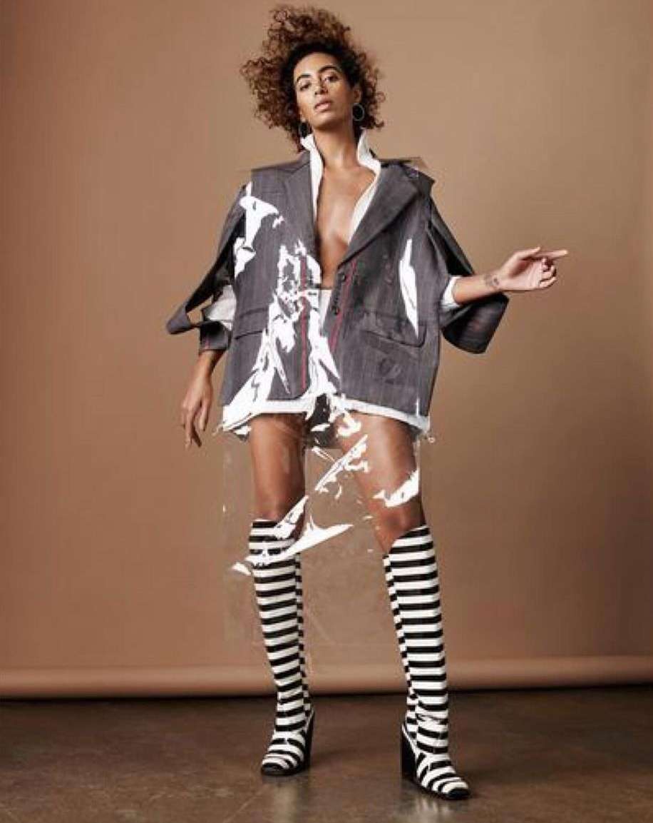 Fashion model posing in blazer, knee-high striped boots, and plastic skirt on a tan background in a studio, pointing to the right.